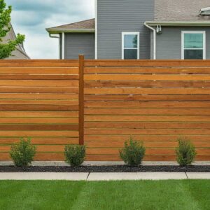 A modern wooden privacy fence with horizontal slats in varying widths, stained a warm brown, encloses a backyard with lush green grass and vibrant flower beds.